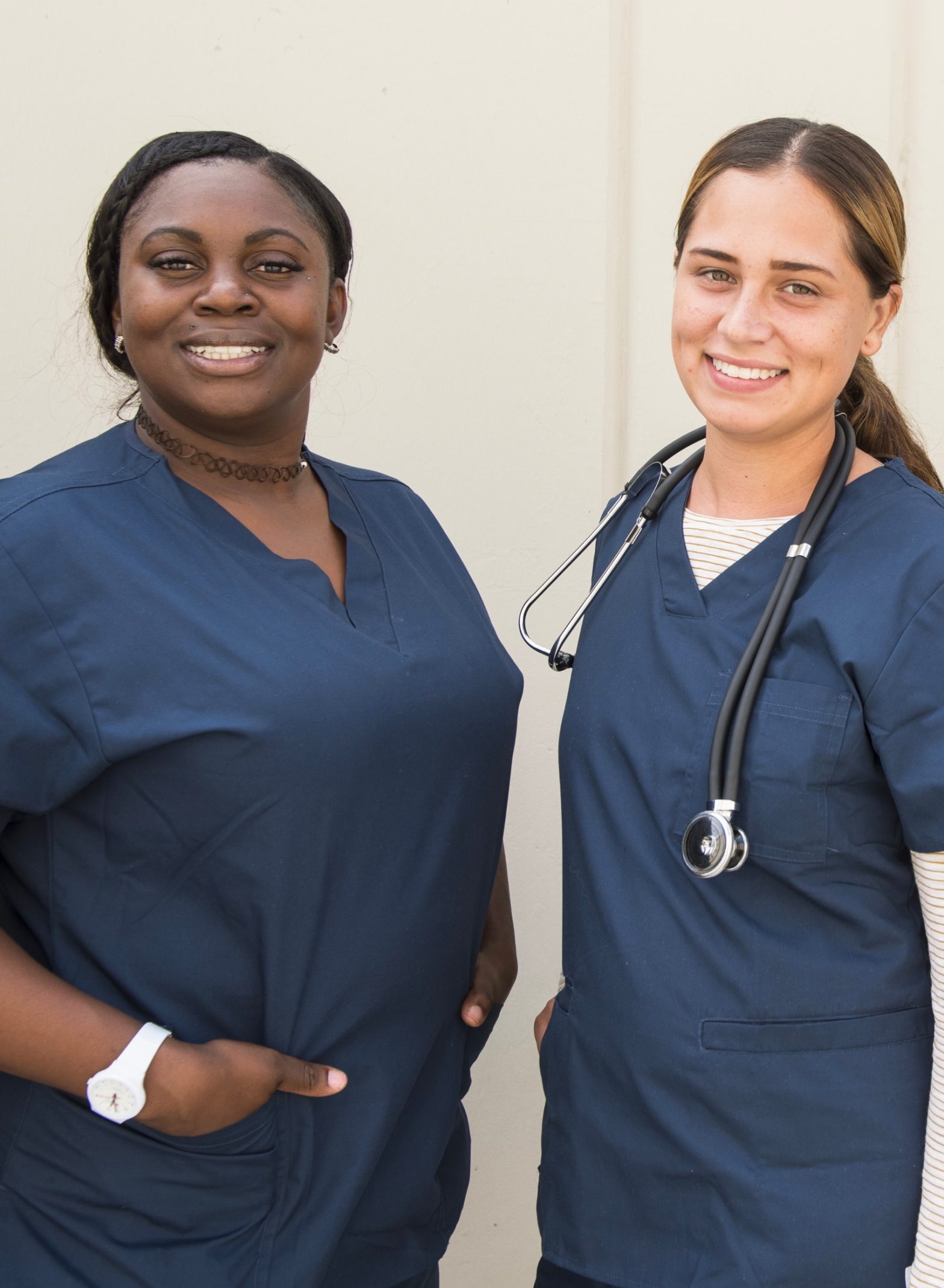 A young black woman and a young white woman, both in navy blue medical scrubs and smiling