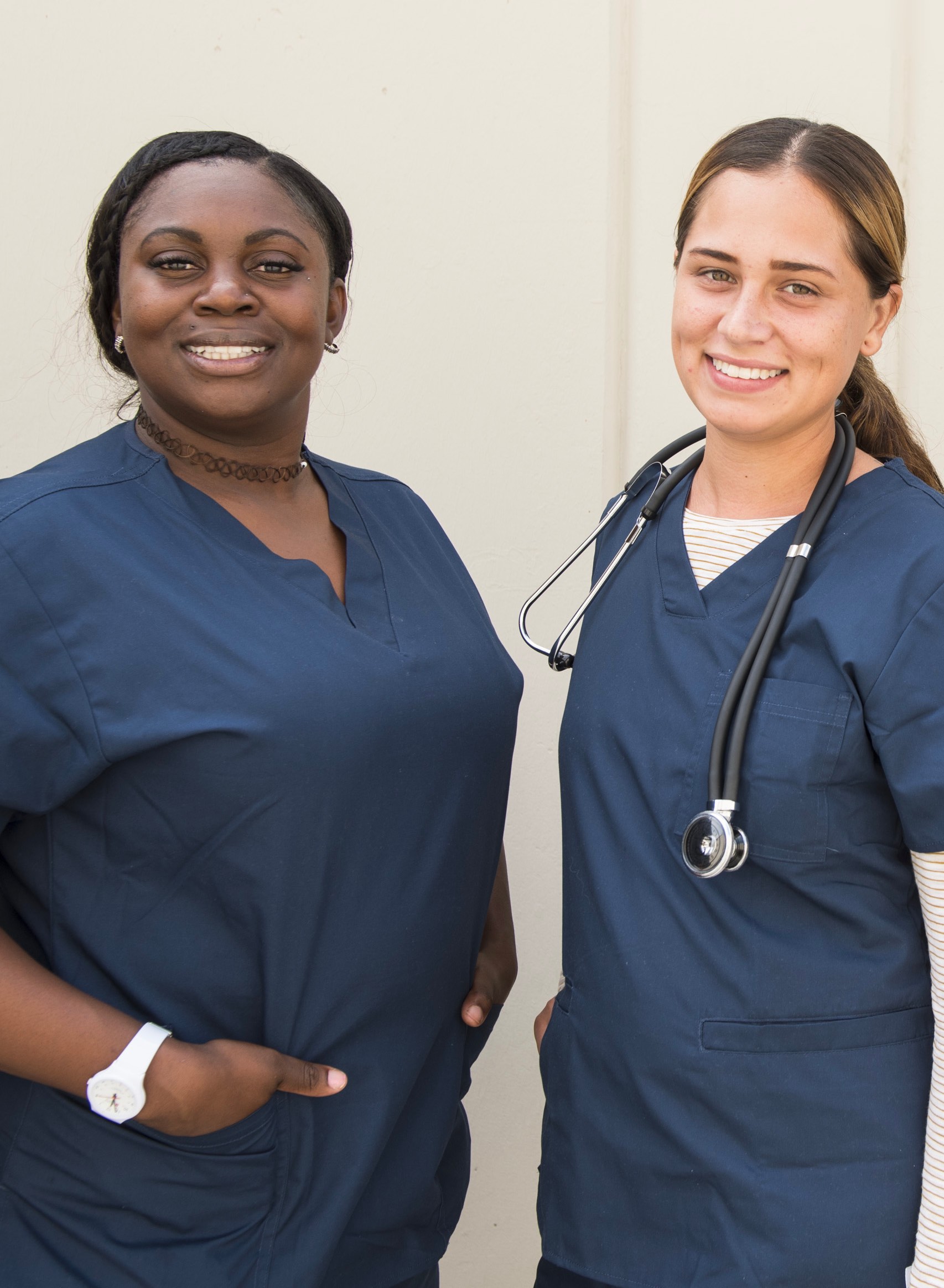 A young black woman and a young white woman, both in navy blue medical scrubs and smiling