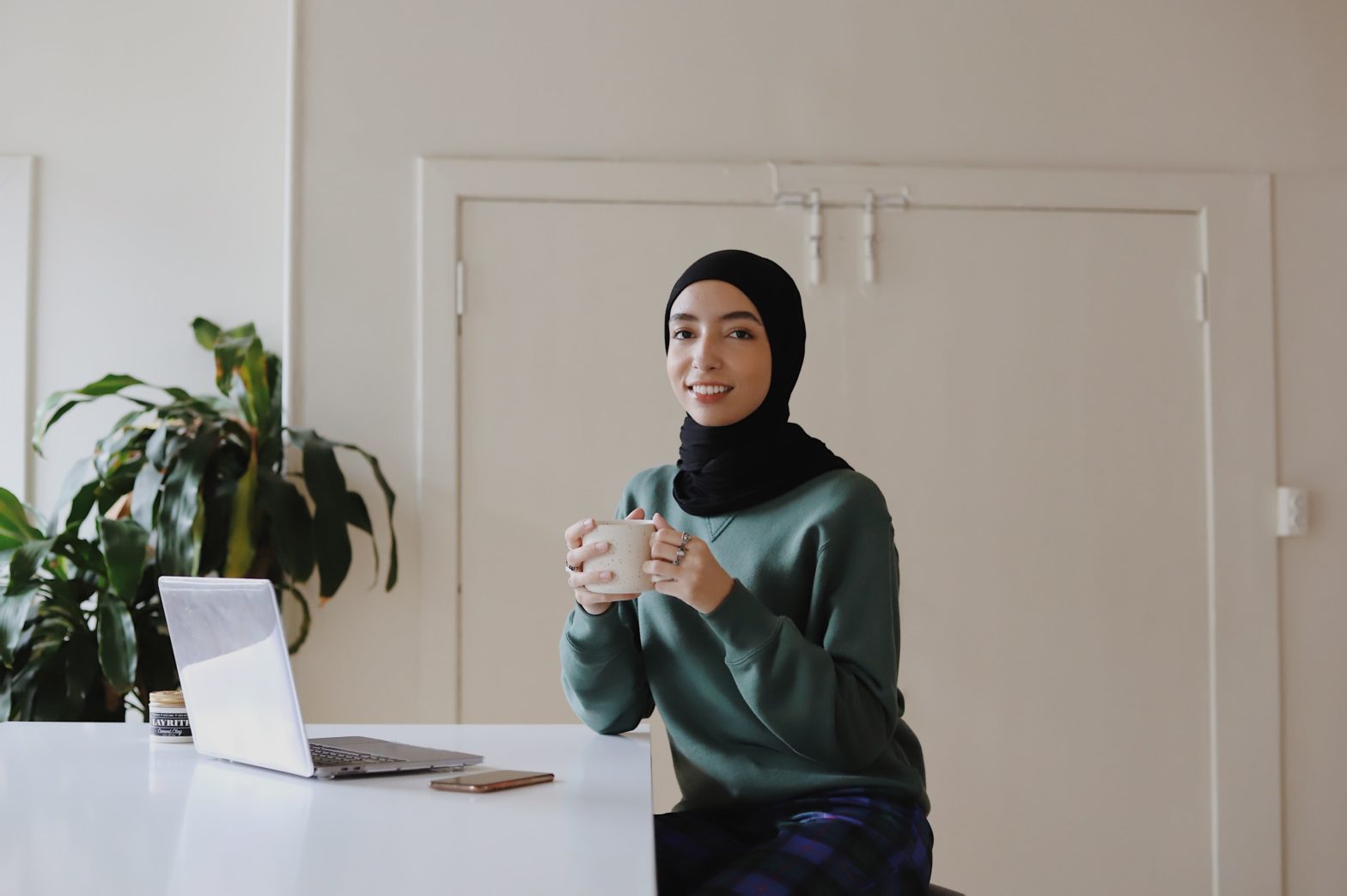 A young woman in a green sweater and a black hijab holding a mug and smiling