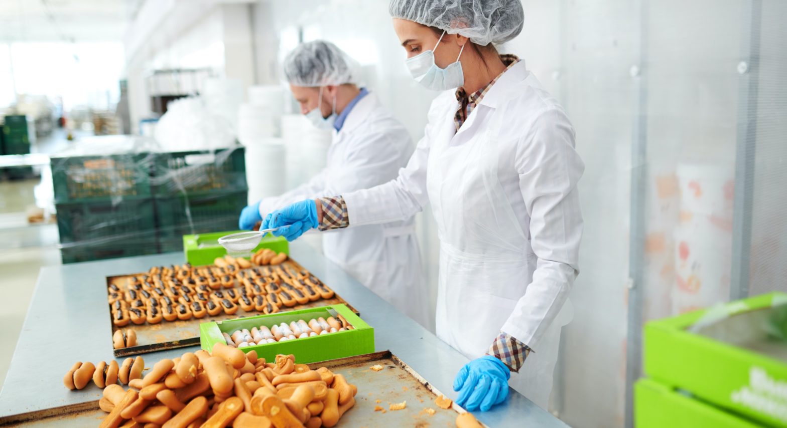 Two food manufacturing employees in white lab coats, gloves, hair nets, and masks, assembling food on an assembly line