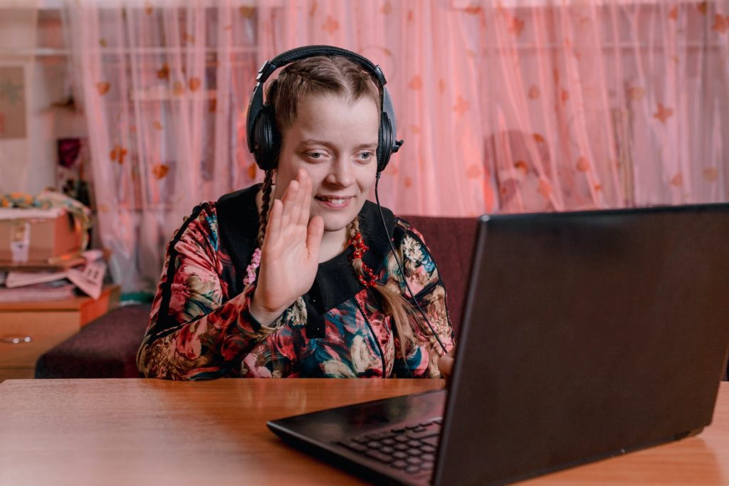 A young person with visual impairment wearing headphones and seated at their laptop, waving to their virtual trainer online